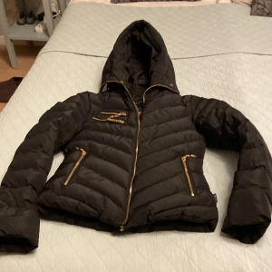 Svart dunjacka från Hollies - Säljer en snygg svart dunjacka från Hollies i storlek 34. Jackan har en cool leopardmönstrad insida och guldfärgade dragkedjor. Perfekt för höst och vinter med sin varma och stiliga design. Den har en huva och långa ärmar, vilket gör den både praktisk och trendig. Det ingår även en päls att sätta på huvan. Jackan är i mycket fint skick och har inga defekter!