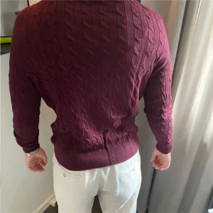 Vinröd stickad tröja ifrån Morris - Säljer en snygg vinröd stickad tröja med kabelstickat mönster. Tröjan har långa ärmar och en normal passform, perfekt för höst och vinter. Den är i bra skick och passar både till vardags och lite finare tillfällen.