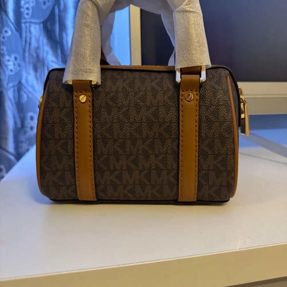 Säljer en snygg brun handväska från Michael Kors ( bedford legacy) i oanvänt  skick. Den är perfekt för både vardag och fest. Kompakt men rymlig nog för dina nödvändigheter!PRIS KAN DISKUTERAS!!!. Väskor.