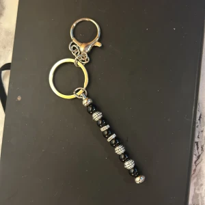 Nyckelring med svarta och silverfärgade pärlor - Snygg nyckelring med en rad av svarta och silverfärgade pärlor. Perfekt accessoar för att ge dina nycklar en stilfull touch. Pärlorna är arrangerade i ett mönster som ger en elegant look. Nyckelringen har en praktisk karbinhake för enkel fastsättning.