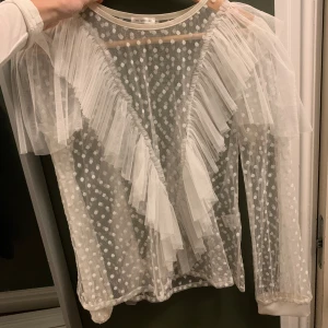 Vit prickig blus från Ida Sjöstedt - Säljer en superfin vit blus från Ida Sjöstedt. Den är i ett transparent material med prickigt mönster och har volangdetaljer framtill. Perfekt för en festlig kväll eller när du vill känna dig extra speciell. Blusen har långa ärmar och en elegant design.