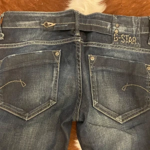G-Star lågmidjade jeans - Säljer ett par snygga G-Star jeans i rå denim. De är mörkblå med en cool tvätt och har en klassisk femficksdesign. Perfekta för både vardag och fest! Passformen är slim och de har en låg midja. Märket är broderat på bakfickan för en extra touch av stil.