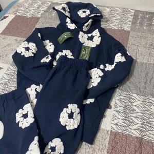 Denim Tears tracksuit - Säljer ett set med en mörkblå hoodie och matchande byxor, båda med stora vita blommor. Hoodien har en klassisk passform med långa ärmar och en bekväm huva. Byxorna har elastisk midja för extra komfort. Perfekt för en avslappnad stil eller mysiga dagar hemma.