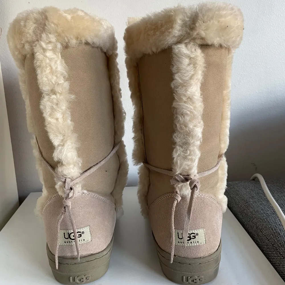 Säljer ett par mysiga höga UGG boots i beige mocka med fluffig pälsdetalj. Perfekta för kalla vinterdagar! De har en bekväm passform och är i mycket bra skick. Snörning baktill för extra stil och komfort.. Skor.