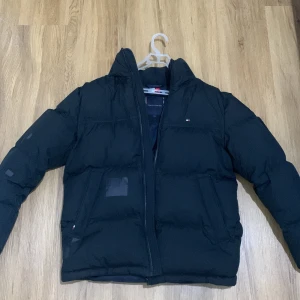 Svart pufferjacka från Tommy Hilfiger - Säljer en svart pufferjacka från Tommy Hilfiger i bra skick. Jackan har en dragkedja framtill och är perfekt för kyliga dagar. Den är långärmad och har en klassisk pufferstil som håller dig varm och bekväm. Perfekt för både höst och vinter! Det har dock hänt saker med jackan men jag fick en grabb och täcka det och det ser underbart ut. Köpte den för 3299