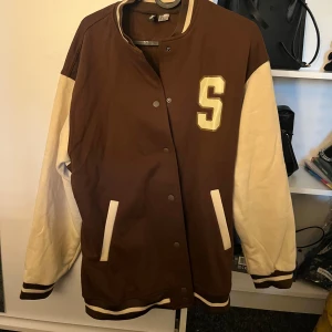 Brun och vit bomberjacka med Seattle-tryck - Säljer en snygg brun och beige ”jacka”. Den har ett stort 'S' på framsidan och 'Seattle' tryckt på baksidan. Jackan har långa ärmar och stängs med knappar. Perfekt för höst och vår! Storlek s 