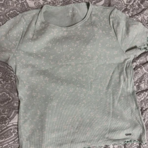 Grön ribbad topp från Hollister - Säljer en söt ljusgrön, pastell grön, ribbad topp från Hollister. Den har korta ärmar och ett mönster med små vita blommor, stretchigt material. Toppen är i mycket bra skick och passar till både jeans och kjol.❤️