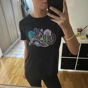 Svart t-shirt från Kenzo - Säljer en svart t-shirt från Kenzo med ett färgglatt blommönster och logga på framsidan. T-shirten är kortärmad och i mycket bra skick. Perfekt för en casual look eller att styla upp med accessoarer.