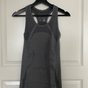 Grå ärmlös träningstopp - Säljer en grå ärmlös träningstopp i syntetmaterial. Perfekt för gymmet eller löprundan. Toppen har en tight passform och meshdetaljer för extra ventilation. Den är i bra skick med en liten slitage på sidan. Perfekt för dig som vill träna med stil!