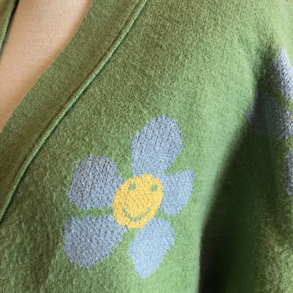 Superfin grön kofta med glada blommor i vitt och gult. Den går ner till höften och har stora vita knappar framtill. Perfekt året om, och ger en härlig retrovibe. Ärmarna är långa och koftan är i mycket bra skick. Mjuk och skön.. Neuletakit & Villapaidat.