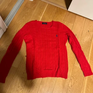 Röd stickad tröja från Ralph Lauren - Säljer en snygg röd stickad tröja från Polo Ralph Lauren. Den har ett klassiskt kabelstickat mönster och en broderad logga på bröstet. Perfekt för höst och vinter, med långa ärmar och en bekväm passform. Passar både till vardags och lite finare tillfällen. Storleken är Xs
