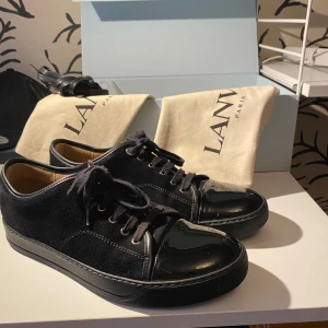 Lanvin skor - Säljer ett par stilrena svarta sneakers från Lanvin i mycket bra skick. Skorna har en snygg kombination av mocka och skinn med glansiga detaljer på tån. Perfekta för både vardag och fest! De har snörning och en bekväm sula. Kommer med originalpåse. Storlek uk8 eller 42-43