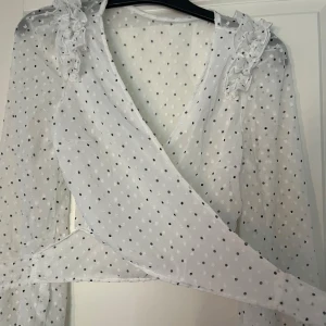 Vit prickig omlottblus - Säljer en superfin vit omlottblus med svarta prickar. Blusen har långa ärmar och volangdetaljer vid axlarna som ger en romantisk touch. Perfekt för både vardag och fest! 💫