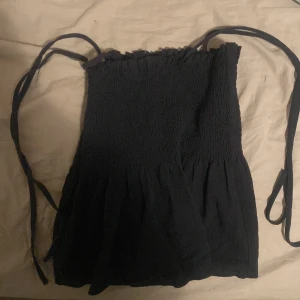 Svart smockad topp med knytband - Säljer en söt svart smockad topp med smala axelband som knyts. Toppen har en elastisk passform och en lätt volangdetalj vid fållen. Perfekt för sommardagar eller en avslappnad kväll ute. Passar bra till jeans eller kjol! !! STORLEK XS !! Bra skick! (Köpt från en annan)