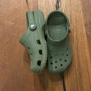 Olivgröna Crocs - Säljer ett par olivgröna Crocs i syntetmaterial. Perfekta för sommarens alla äventyr med sin bekväma passform och ventilerande hål. De har en justerbar hälrem för extra stöd. Använd dem på stranden eller i trädgården för maximal komfort.