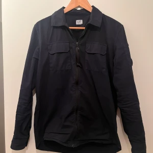 Svart overshirt från C.P. Company - Säljer en stilren svart overshirt från C.P. Company i mycket bra skick. Den har en dragkedja framtill och två bröstfickor med lock. Perfekt för höst och vår, och funkar både till vardags och fest. Passar bra över en t-shirt eller tröja. Den är köpt på Nääs fabrik för runt 4500 men jag säljer för ändast 999 kr, kvitto finns tyvärr ej kvar 