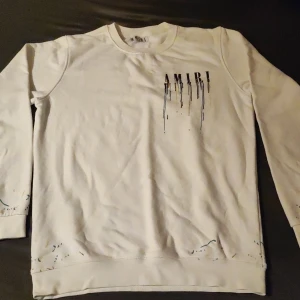 Vit tröja från Amiri med färgstänk - Säljer en snygg vit tröja från Amiri med coola färgstänk och droppande logotyp. Perfekt för en avslappnad men stilren look. Tröjan har långa ärmar och är i bra skick. Passar perfekt till både vardag och fest!
