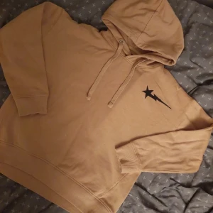Beige hoodie med tryck - Säljer en snygg beige hoodie med en cool svart stjärna på bröstet. Den har en bekväm passform och är perfekt för både höst och vår. Huvan har dragsko och ärmarna är långa med ribbade muddar. Perfekt för en avslappnad stil!
