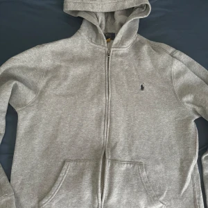 Grå hoodie från Ralph Lauren - Säljer en snygg grå hoodie från Ralph Lauren i storlek Xs. Den har en dragkedja framtill och en klassisk broderad logga på bröstet. Perfekt för både höst och vinter, med en mysig insida som håller dig varm. Passar lika bra till vardags som till träning. 🐴, priset kan även diskuteras. 