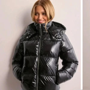 Svart glansig pufferjacka - Säljer en supercool svart pufferjacka med glansigt material. Den har en dragkedja framtill och en mysig huva. Perfekt för kalla dagar och ger en snygg look. Jackan har en normal passform. Har ett litet hål, skriv för fler bilder💕