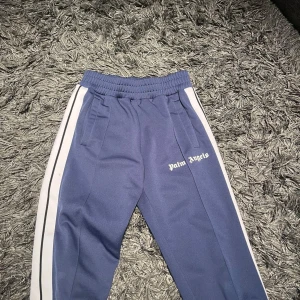 Blå träningsbyxor från Palm Angels - Säljer ett par snygga blå träningsbyxor från Palm Angels i storlek S. Byxorna har vita sidoremsor och elastisk midja för extra komfort. Perfekta för både träning och chill. De är i mycket bra skick och redo för nya äventyr! 💙