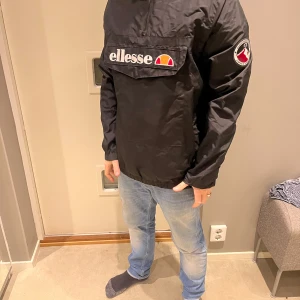 Svart jacka från Ellesse - Säljer en svart jacka från Ellesse i mycket bra skick. Den har en stor logga på bröstet och en cool patch på ärmen. Jackan är perfekt för höst och vår, med en normal passform och långa ärmar. Den är gjord av syntetmaterial och har en dragkedja framtill. Perfekt för vardagsbruk!