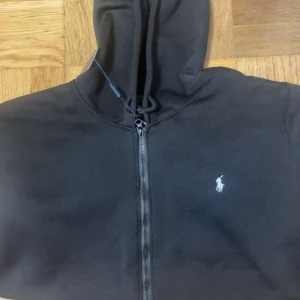 Svart hoodie från Ralph Lauren - Säljer en stilren svart hoodie från Ralph Lauren med dragkedja och klassisk logga på bröstet. Perfekt för höst och vinter, med en skön passform och fickor framtill. Huvan ger extra värme och stil. Passar både till vardags och chillkvällar. 🖤