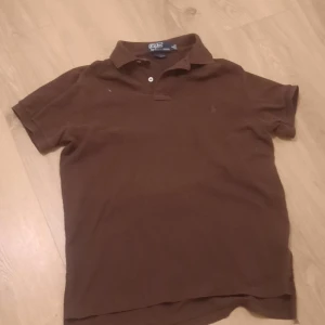 Brun pikétröja från Polo - Säljer en klassisk brun pikétröja från Polo i bra skick. Den har korta ärmar och en stilren krage med två knappar. Perfekt för en avslappnad men ändå snygg look. Passar bra till både jeans och shorts!