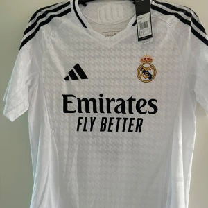 Vit Real Madrid fotbollströja Mbappe  - Säljer en vit Real Madrid fotbollströja i storlek L från Adidas. Tröjan har korta ärmar och är i nytt skick. Den har klubbens emblem och sponsorlogga på framsidan samt spelarnamnet 'Mbappé' och nummer 9 på baksidan. Storlek L