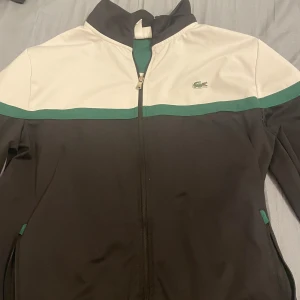 Svart och vit jacka från Lacoste - Säljer en snygg svart och vit jacka från Lacoste i storlek L. Jackan har en dragkedja framtill och en grön rand som ger den en sportig look. Perfekt för höst och vår! 🐊