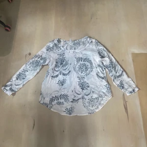Vit blus från Soyaconcept - Säljer en fin vit blus från Soyaconcept med ett vackert blommigt mönster i grått. Blusen har långa ärmar och en rund halsringning. Perfekt för både vardag och festliga tillfällen!
