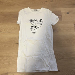 Vit t-shirt klänning med konstnärligt ansikte - Säljer en vit t-shirt klänning med ett coolt konstnärligt ansikte tryckt på framsidan. Den är kortärmad och i bomull, perfekt för en avslappnad stil. Storleken är normal och den är i mycket bra skick.