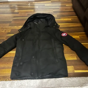 Svart vinterjacka från Canada Goose - Säljer en svart vinterjacka från Canada Goose i mycket bra skick. Jackan har en klassisk design med långärmade ärmar och en praktisk huva. Perfekt för kalla vinterdagar. Den har en broderad logotyp på ärmen och stängs med dragkedja och knappar. Supervarm och stilren! Passar även till storlek M.