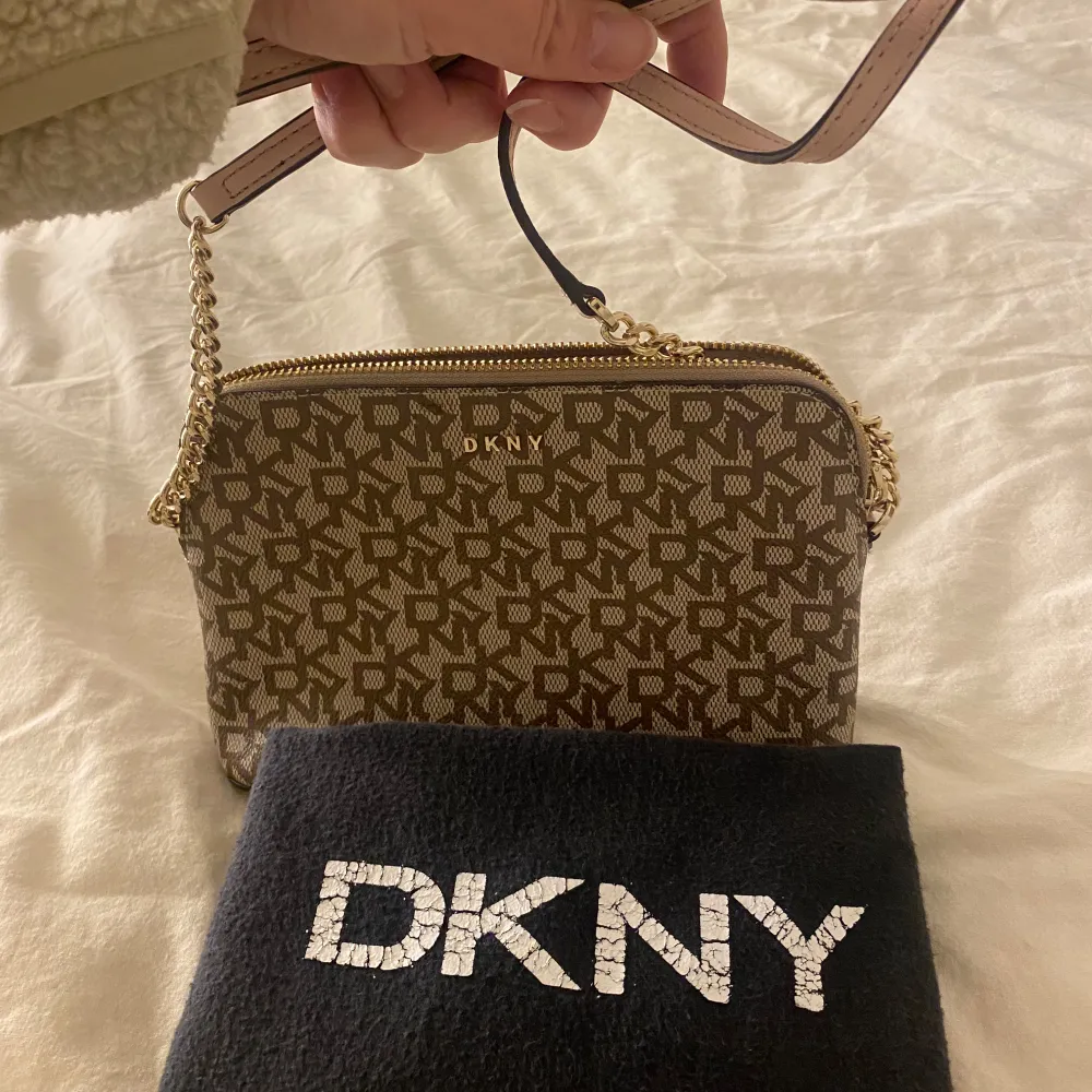 Säljer en snygg brun axelväska från DKNY med deras klassiska logotypmönster. Väskan har en guldfärgad dragkedja och kedjedetaljer på axelremmen. Perfekt för både vardag och fest! 👜✨. Väskor.