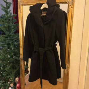 Svart kappa med luva  - Säljer en elegant svart kappa med huva och knytskärp i midjan. Perfekt för höst och vinter. Den har långa ärmar och en stilren design som passar både till vardags och fest. 