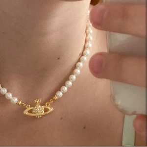 Pärlhalsband med guldberlock från Vivienne Westwood - Säljer ett pärlhalsband från Vivienne Westwood med en ikonisk guldberlock i form av en planet. Halsbandet har vita pärlor och en fin guldfärgad låsdetalj. Perfekt för att ge något extra till vilken outfit som helst. Passar både till vardag och fest!