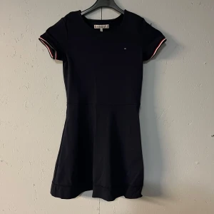 Mörkblå klänning från Tommy Hilfiger stl 140 - Snygg mörkblå klänning från Tommy Hilfiger i ett klassiskt snitt. Den har korta ärmar med vita och röda detaljer vid ärmsluten. Perfekt för både vardag och festliga tillfällen. Materialet känns mjukt och bekvämt. En tidlös design som passar alla säsonger.