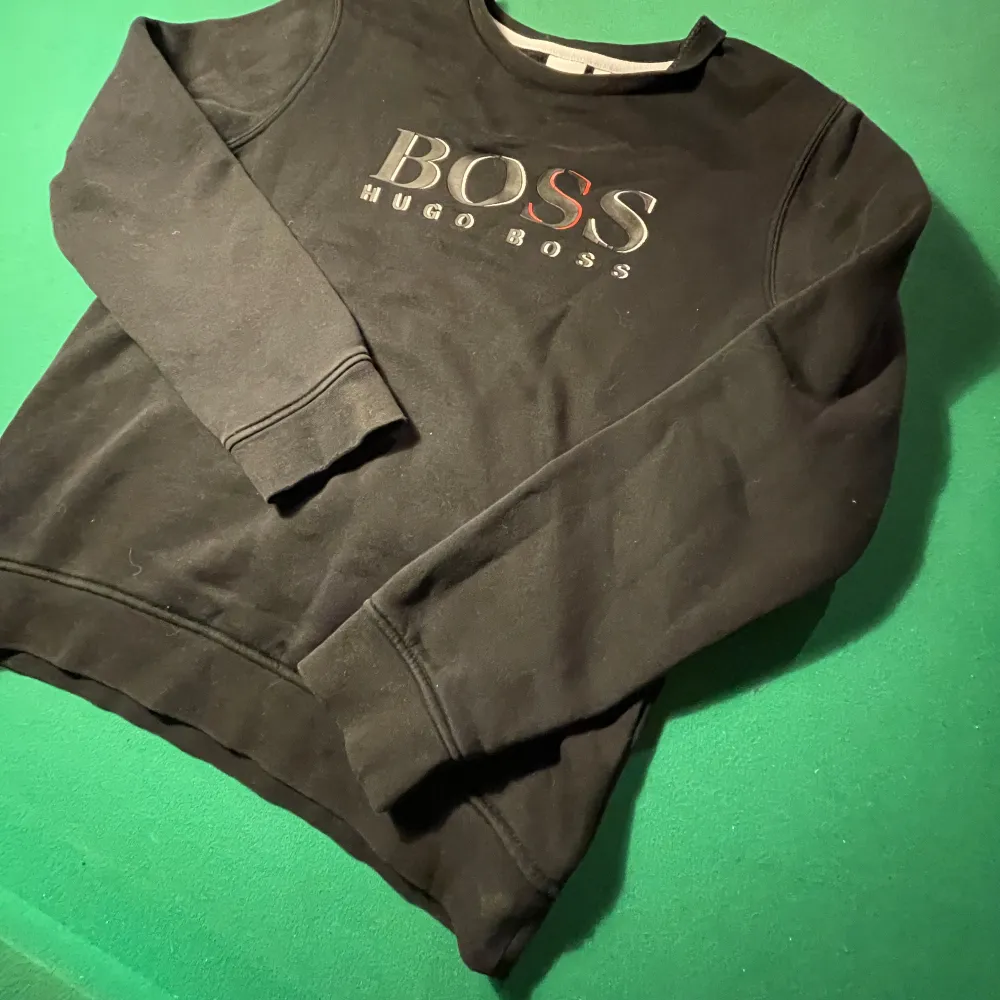 Säljer en stilren svart tröja från Hugo Boss i storlek 160. Tröjan har långa ärmar och ett klassiskt tryck med BOSS-loggan på bröstet. Perfekt för både vardag och festliga tillfällen. Skön och enkel att matcha med det mesta i garderoben! Priset är verkligen inte hugget i sten!. Tröjor & Koftor.