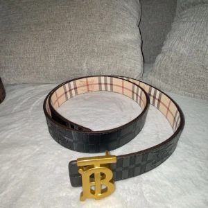 Burberry - Säljer ett stilrent burberry skärp med ett rutigt mönster i svart och beige. Perfekt för att ge en lyxig touch till din outfit. Passar både till vardag och fest! Obs:skriv innan köp!