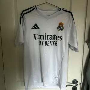 Säljer en vit Real Madrid fotbollströja från Adidas i mycket bra skick. Tröjan har korta ärmar och klubbens emblem på bröstet. Den är gjord i ett lätt och andningsbart material, perfekt för både träning och match. Klassisk design med tre svarta ränder på axlarna. Passar perfekt för alla Real Madrid-fans! ⚽️