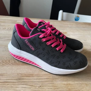 Svarta sneakers med rosa detaljer - Snygga svarta sneakers med rosa snörning och innersula. Skorna har en vit sula med rosa detaljer på sidan. Perfekta för en casual look eller träning. De har ett mönster av olika symboler på ovansidan som ger en unik touch.Helt nya 