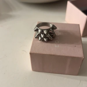Silvrig ring med nitar från Edblad - Cool och edgy ring i silver från Edblad. Den har flera små nitar som ger en tuff look. Perfekt för att ge din outfit en extra edge. Ringen är i mycket bra skick och kommer i originalförpackning. Strl 16,80! Köpt för 399 säljer för 150 kr pris kan diskuteras