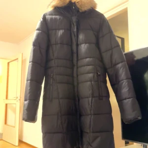Svart lång dunjacka med pälsdetalj - Säljer en svart lång dunjacka med mysig pälsdetalj på huvan. Jackan har en quiltad design och dragkedja framtill. Perfekt för kalla vinterdagar! 🖤 aldrig använt väldigt bra skick