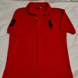 Röd pikétröja från Polo - Säljer en snygg röd pikétröja från Polo i storlek L. Tröjan är i 100% bomull och har en klassisk krage med knappar. Den har en mörkblå broderad logga på bröstet och siffran 3 på ärmen. Perfekt för en avslappnad men stilren look!