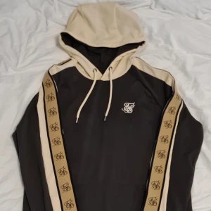 Svart och beige hoodie från SikSilk - Säljer en snygg svart hoodie från SikSilk med beige detaljer och logga på ärmarna. Den har en justerbar huva med dragsko och är perfekt för en avslappnad stil. Passar bra till både vardag och träning. 🖤