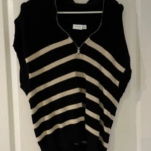 Svart och beige väst från Vila - Snygg svart väst med beige ränder från Vila. Den har en dragkedja framtill och är perfekt för lager-på-lager-stil. Passar bra till både vardag och festliga tillfällen.