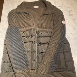 Moncler cardigan - Säljer en snygg grå stickad jacka från Moncler. Den har en quiltad design på framsidan och ärmarna är långärmade. Jackan har en dragkedja framtill och två fickor med knappar. Perfekt för höst och vår!