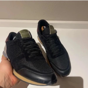 Svarta sneakers från Valentino - Snygga svarta Valentino sneakers i mycket bra skick. De har en stilren design med svarta snören och en diskret logga på plösen. Skorna har en bekväm sula i beige och svart, perfekt för både vardag och fest. En touch av mörkröd detalj på hälen ger dem en unik look. Storlek 43,5