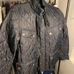 Svart quiltad jacka från Barbour - Säljer en snygg svart quiltad jacka från Barbour. Den har en stilren design med knappar och dragkedja framtill. Jackan har flera praktiska fickor och en cool illustration på insidan.  Jag säljer denna jacka för att den är för liten, nypris 2400kr, pris kan diskuteras!