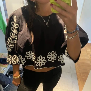 Svart blus med blommigt mönster - Säljer en superfin svart blus med vita blommor broderade längs ärmarna och nederkanten. Blusen är croppad och har trekvartsärmar, vilket gör den perfekt för både vår och sommar. Den är i mycket bra skick!